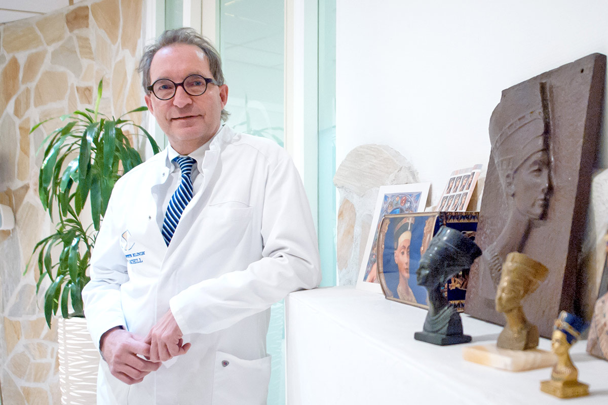 Willkommen in der Nofretete-Klinik - Ein Mann in einem weien Kittel und mit Brille steht neben einem Regal mit Statuen.